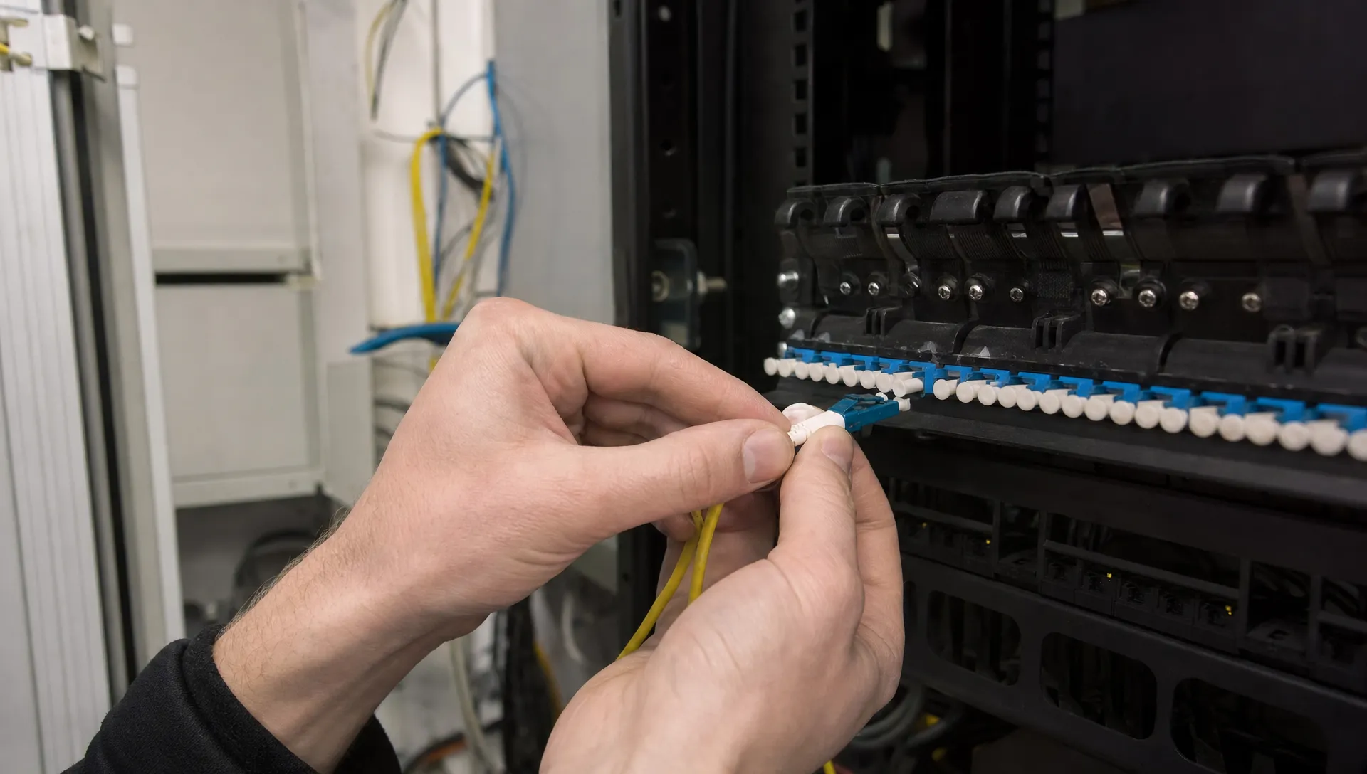 Close-up van handen die een glasvezelconnector plaatsen in een patchpaneel in een kleine technische ruimte voor een verbinding tussen locaties.