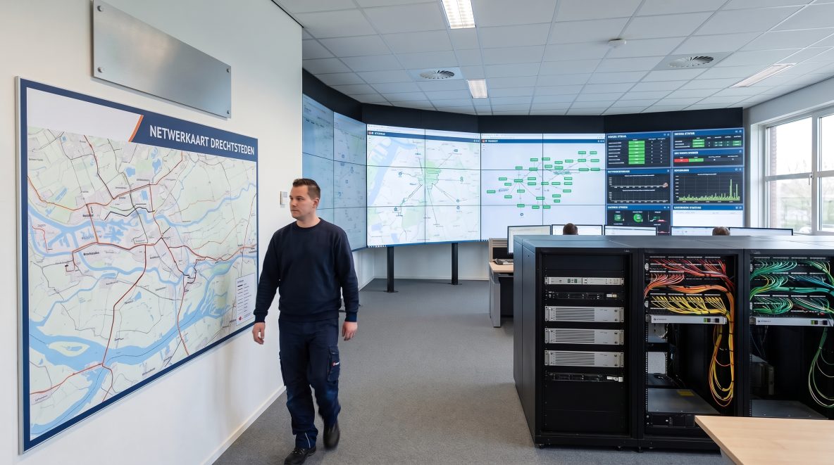Een netwerkoperatieruimte met racks, patchpanelen, schermen en een wandkaart van verbindingen tussen locaties.