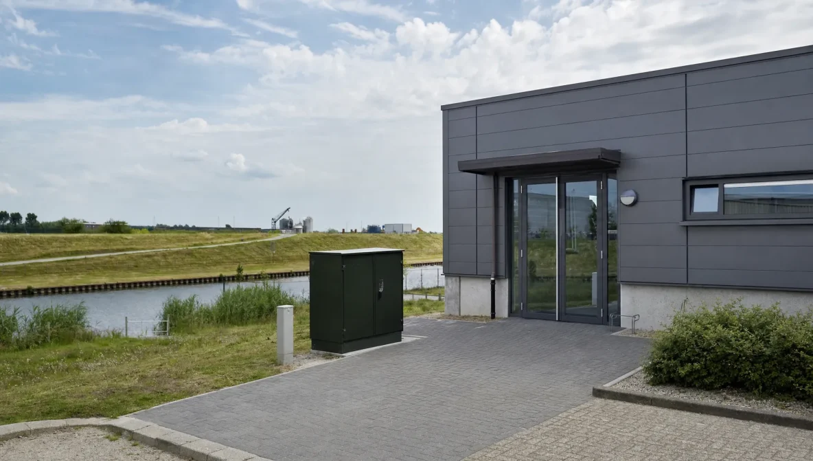 Een technische locatie aan de rand van een bedrijventerrein met een glasvezelkast, een kanaal en een dijk in het landschap.